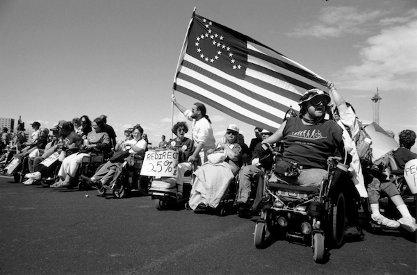 A large group of disability activists in wheelchairs outside in the city. Many holding signs and a man holding a large flag